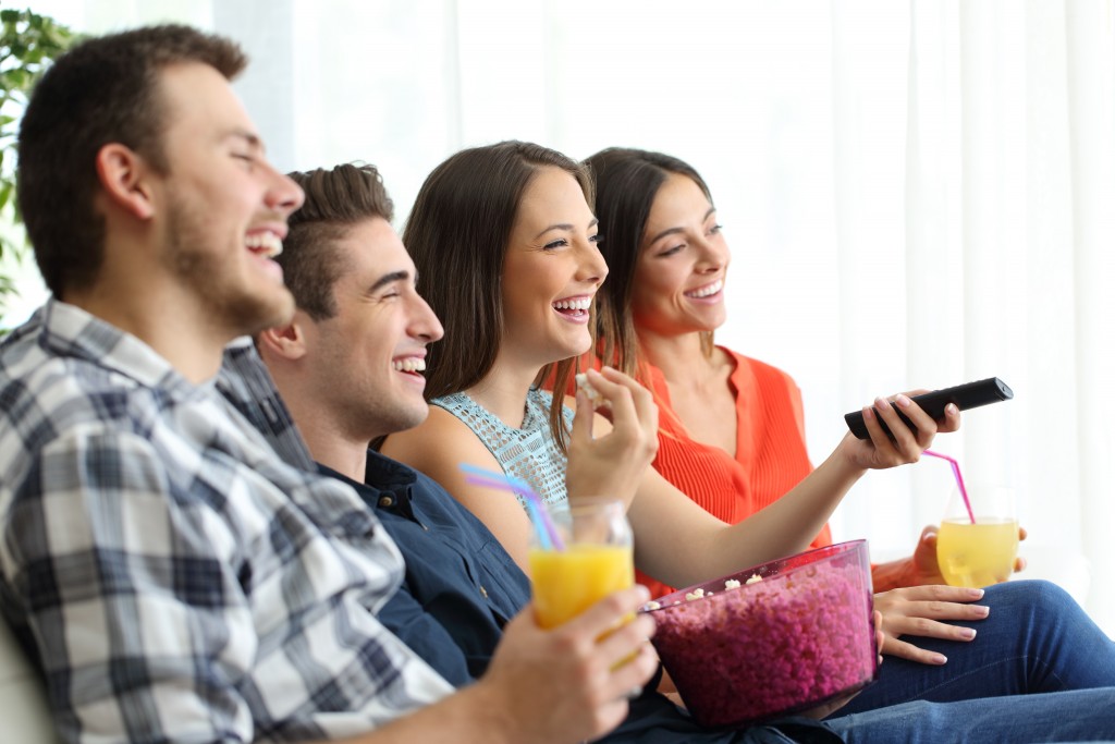 Side view of a happy group of four friends watching and enjoying tv sitting on a sofa in the living room at home